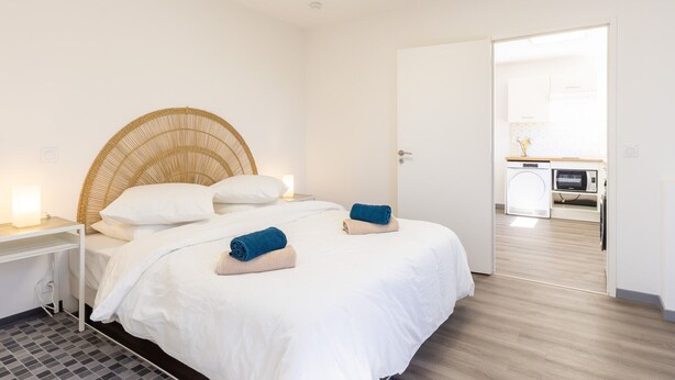 Een slaapkamer met een bed, nachtkastje en uitzicht op de keuken.