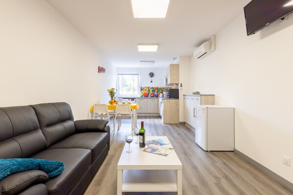 A modern living room with a black leather sofa, a white coffee table with a wine bottle and glasses, and a colorful painting. A kitchen with white cabinets and a white refrigerator is visible in the background.
