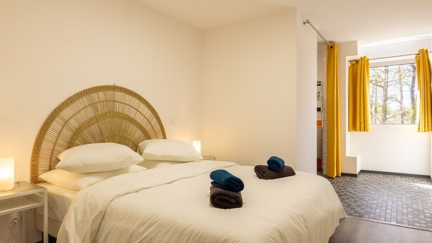 A hotel room with a bed, a nightstand, a wicker headboard, and a window with yellow curtains.
