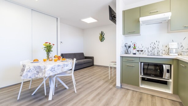 Een moderne keuken met een ingebouwde oven, een eethoek met een tafel voor twee en een grijze bank in de woonkamer.