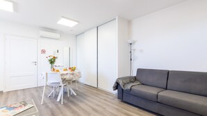 Een moderne woonkamer met een grijze bank, een eettafel met stoelen, een witte muur en een deur met een 'Niet roken'-bordje.