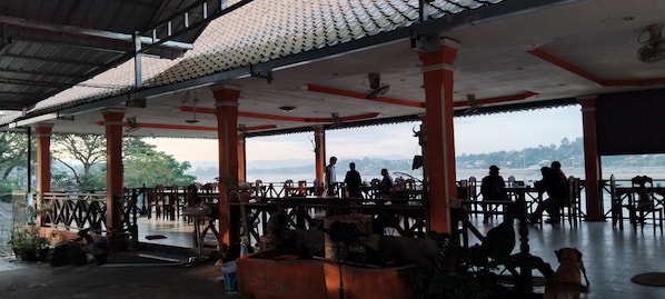 An outdoor dining area with multiple tables and chairs, a tiled roof, and a view of a body of water.