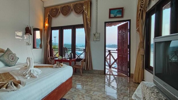 A hotel room with a bed, a table, a chair, a TV, and a balcony with a view. - Houayxai Riverside Phonevichith Hotel (Houayxay)