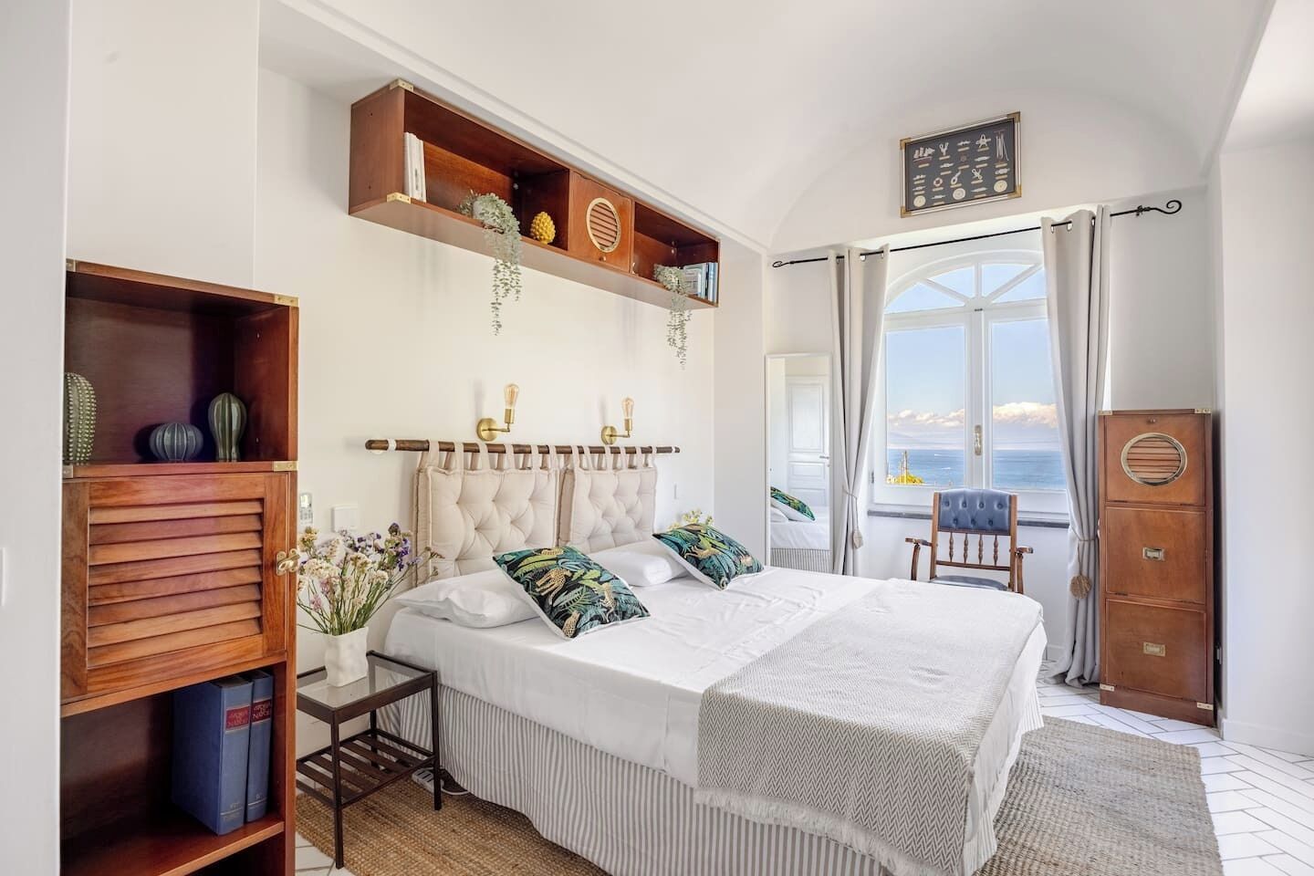Un dormitorio con una cama grande, un cabecero de madera, una mesita de noche con libros, una ventana con cortinas y vistas al mar.