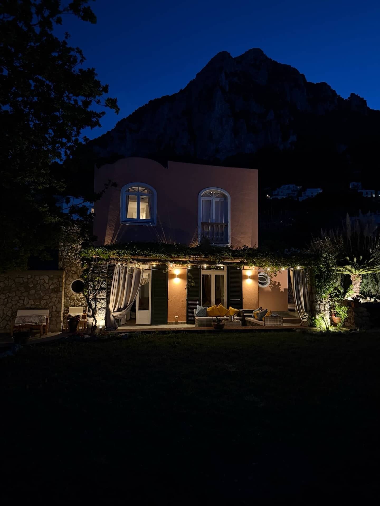 A two-story house with a mountain backdrop, featuring a patio with outdoor seating and string lights.