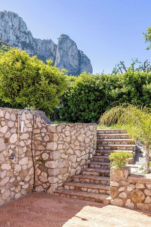 Una escalera de piedra que conduce a un muro de piedra con ducha, rodeada de follaje y un paisaje montañoso.