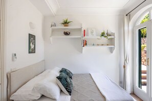 A bedroom with a bed, pillows, a bedside table, a wall-mounted shelf with books and decorative items, and a view through an open door.