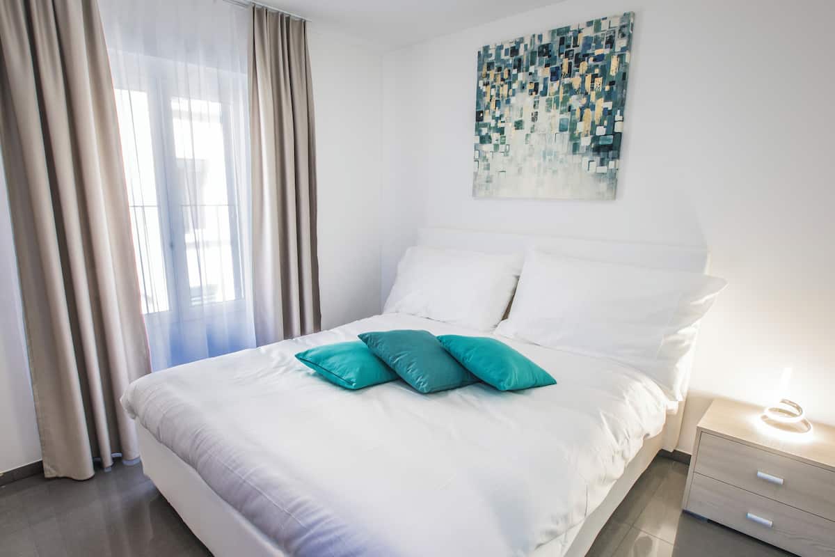A neatly made bed with white and teal pillows, a modern abstract painting on the wall, and a bedside table with a lamp.