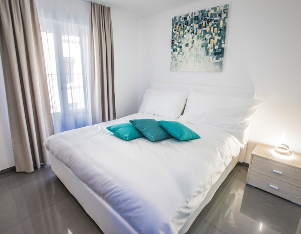Cama bem arrumada com almofadas brancas e azul-turquesa, pintura abstrata moderna na parede e mesa de cabeceira com abajur.