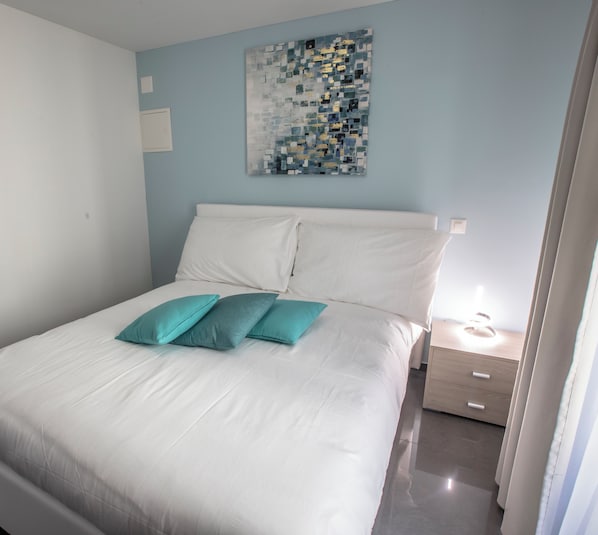 Un letto rifatto con lenzuola bianche e cuscini color acquamarina, un piccolo comodino con una lampada e un'opera d'arte appesa sopra il letto.