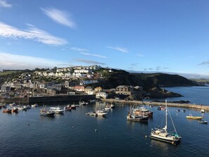 A coastal town with a harbor filled with boats, houses on a hillside, and a lighthouse in the distance. - Orchard View (St Austell)