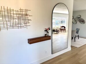 A modern interior with a large mirror, a geometric wall art piece, a wooden shelf, and a view of a living room through the mirror. - Luxury Modern Waterfront Villa - Saltwater Pool / Fishing Dock / BBQ - Sleeps 12 (Tampa)