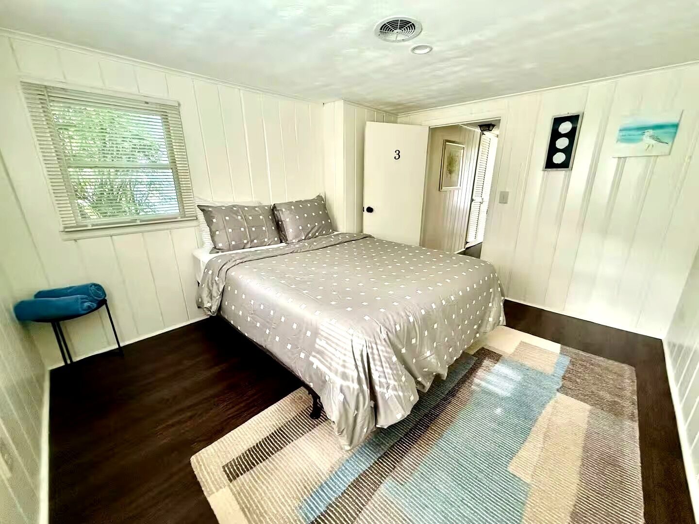 Ein Schlafzimmer mit Bett, Fenster mit Jalousien, einer Tür mit der Nummer 3 und einem Wandbild.