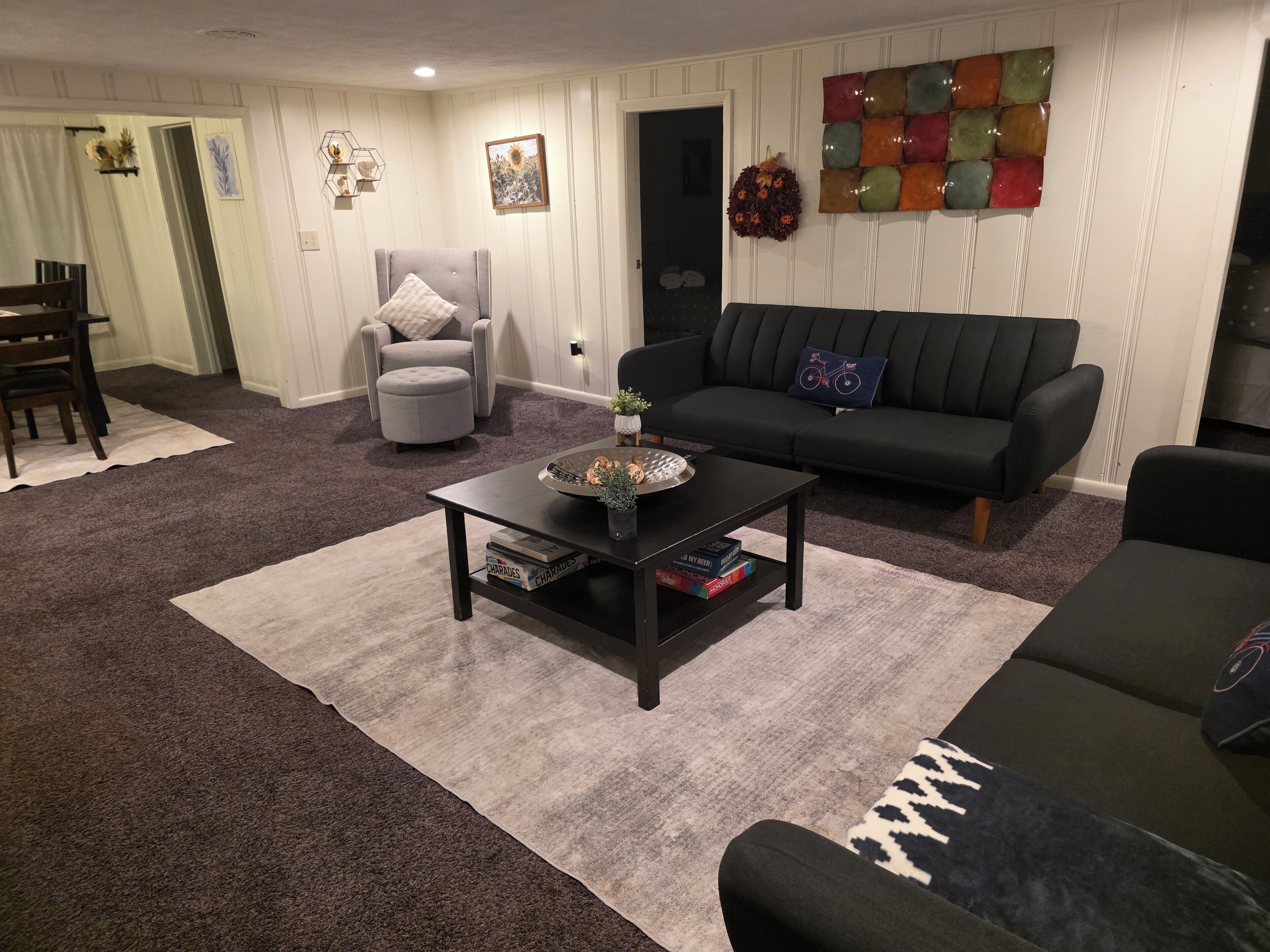 Uma sala de estar com um sofá escuro, uma poltrona cinza, uma mesa de centro com livros e um tapete.