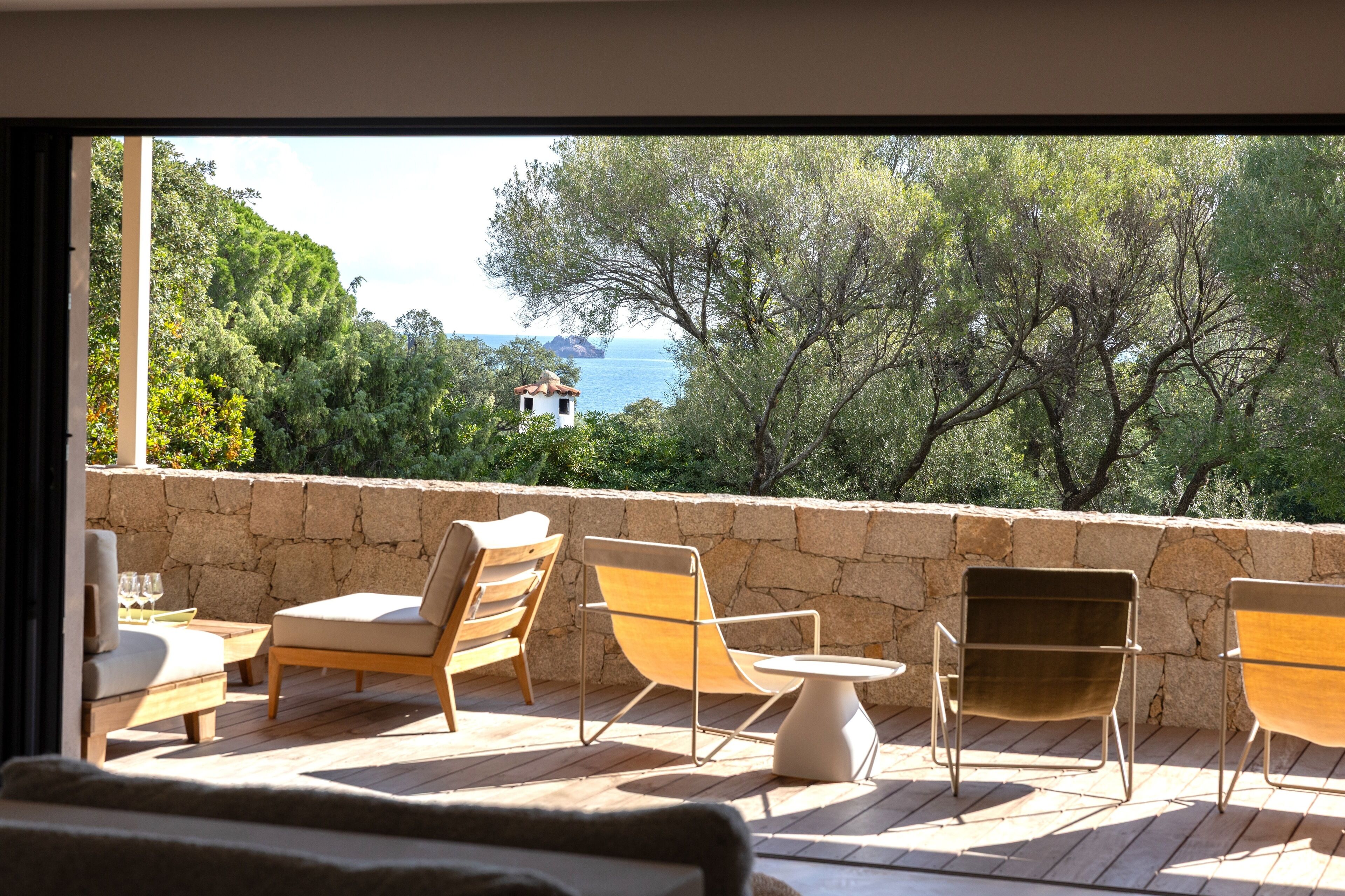 A balcony with wooden flooring, a stone wall, and a view of the sea and trees.