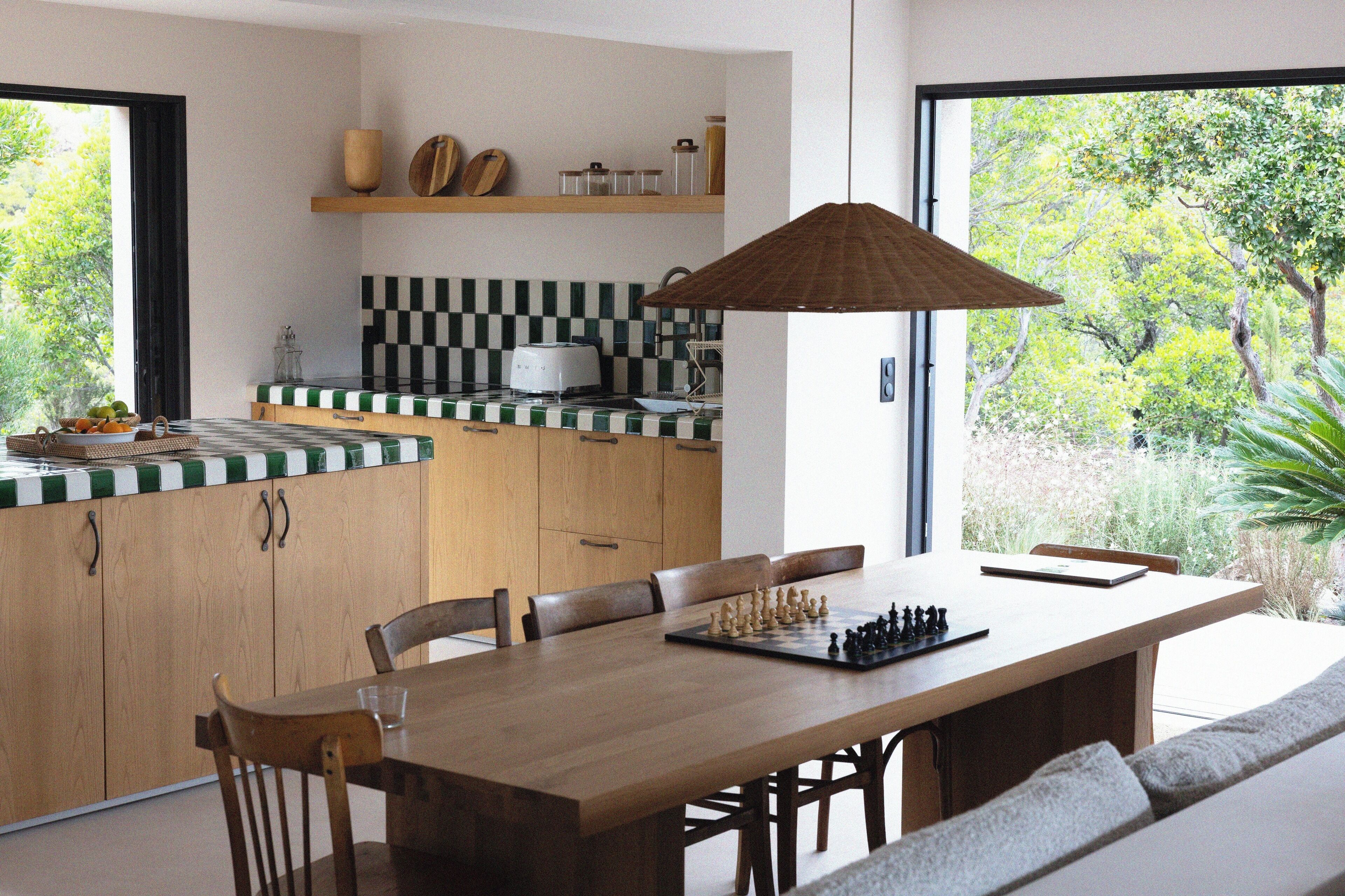 Een moderne keuken met een houten tafel en stoelen, een schaakbord en een groot raam met uitzicht op groen.