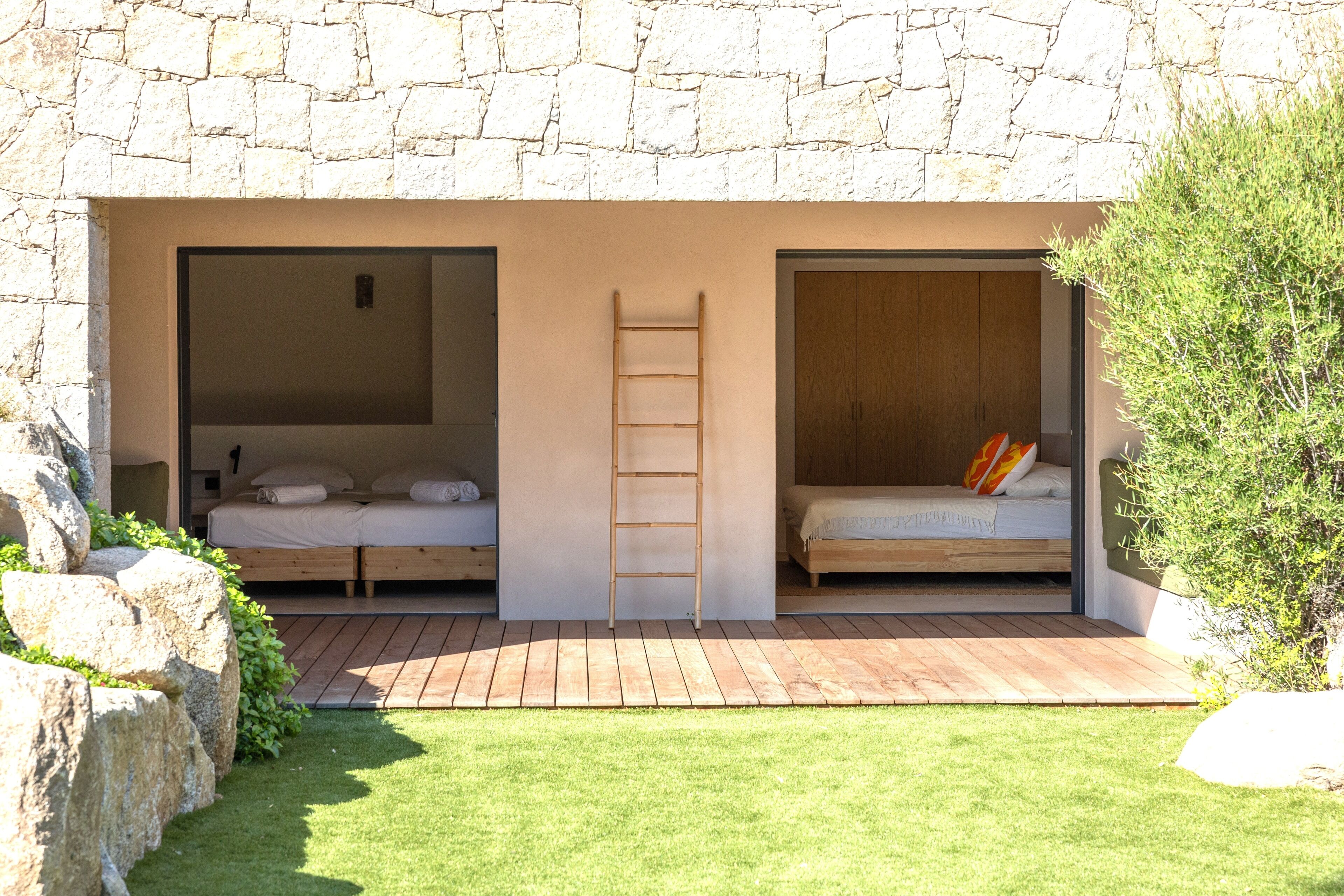 A modern bedroom with a stone wall, two beds, a wooden ladder, and a small outdoor area with grass and rocks.