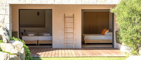 A modern bedroom with a stone wall, two beds, a wooden ladder, and a small outdoor area with grass and rocks.