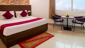 Habitación de hotel con una cama grande, cabecera de madera, manta roja, una mesita con frutero, dos sillas, ventana con vistas y ventilador de techo.