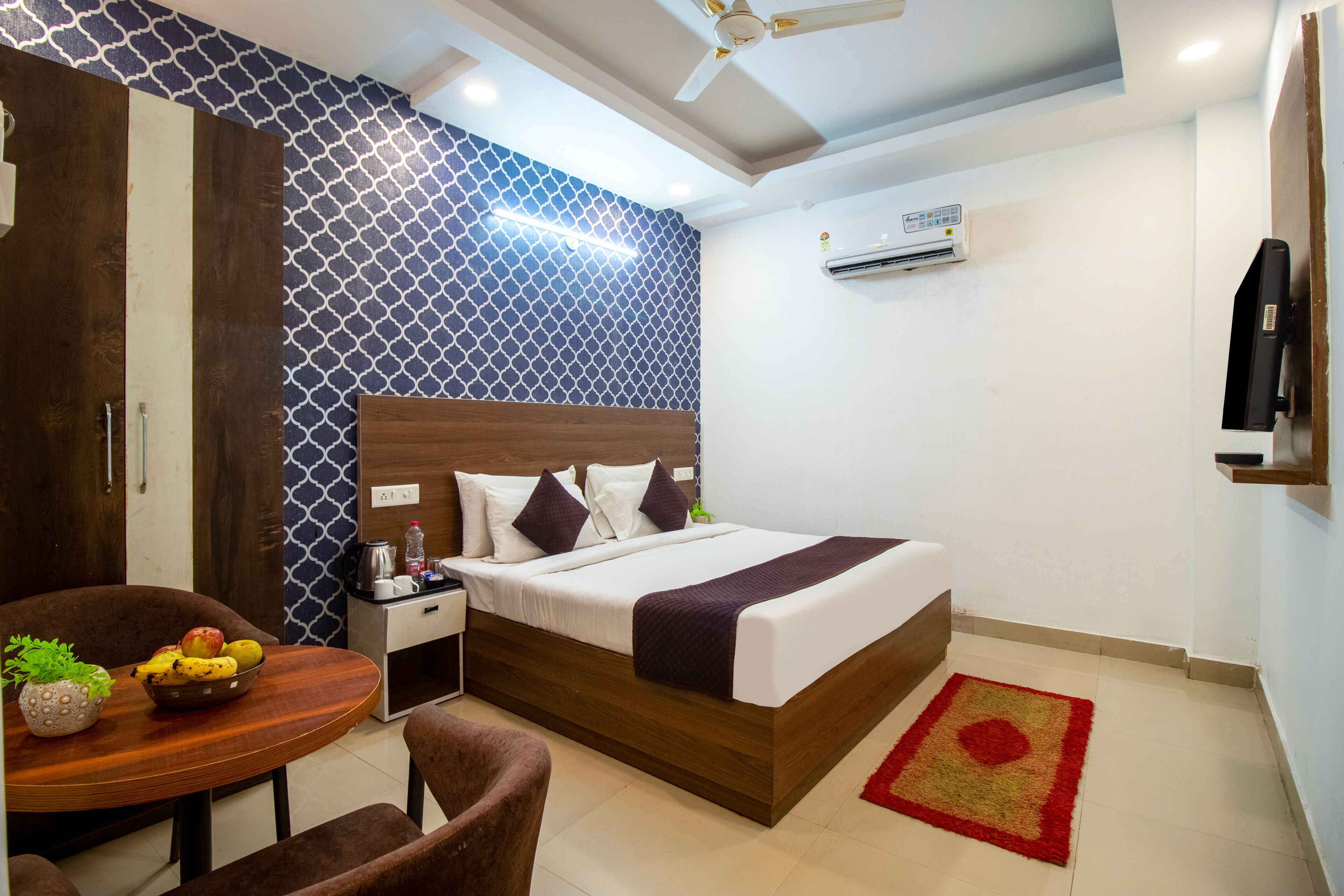 A hotel room with a bed, a desk with a chair, a dining table with fruit, and a red rug.
