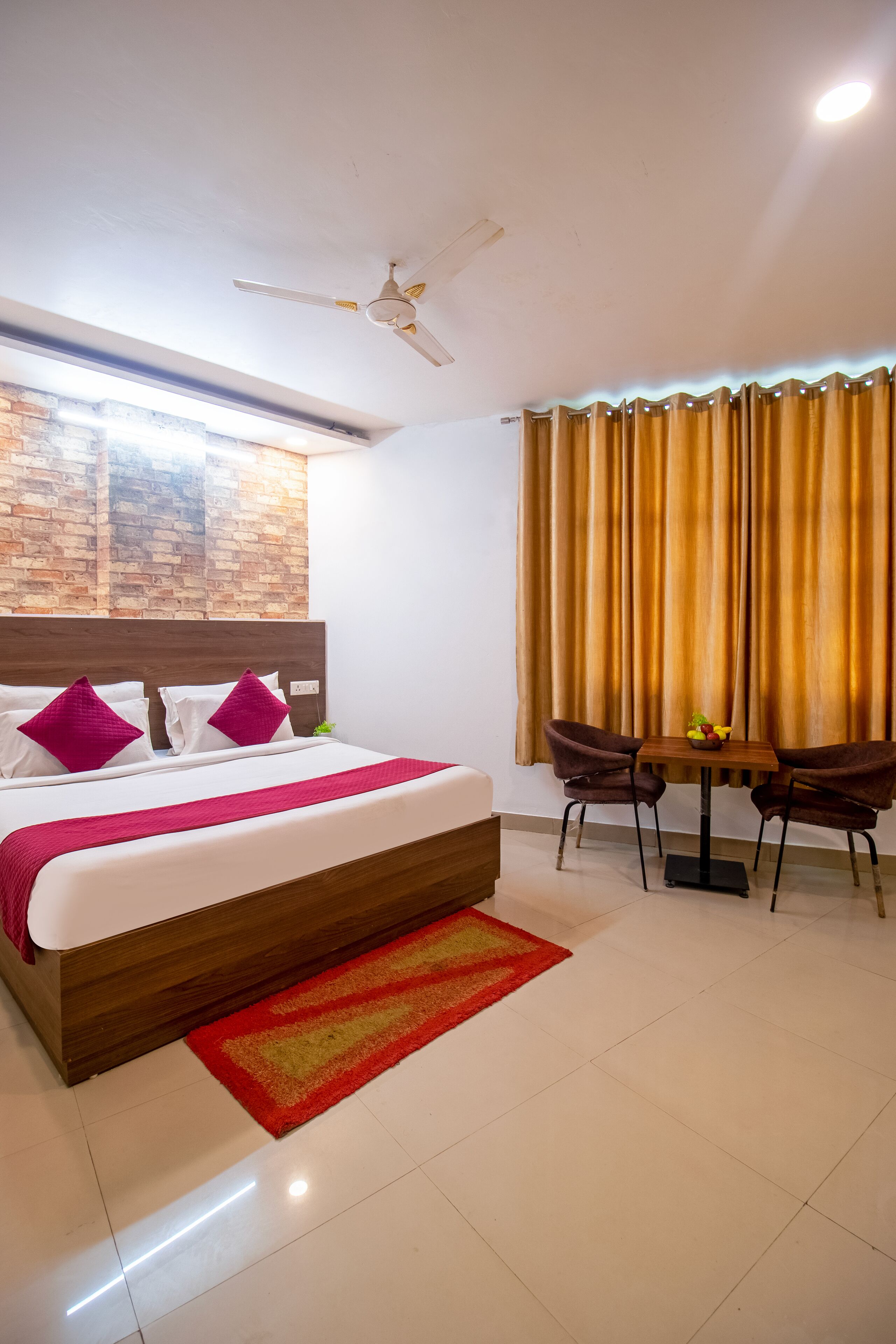 A hotel room with a large bed, a wooden headboard, a red and yellow striped rug, a ceiling fan, a table with two chairs, and a window with curtains.