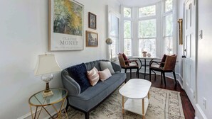 Uma sala de estar com um sofá cinzento, uma mesa de centro de vidro e um espaço de refeições com cadeiras de vime e uma mesa pequena. Há obras de arte emolduradas nas paredes, incluindo uma de Claude Monet.