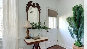 Um corredor com porta branca, espelho, mesa com abajur e planta em vaso.
