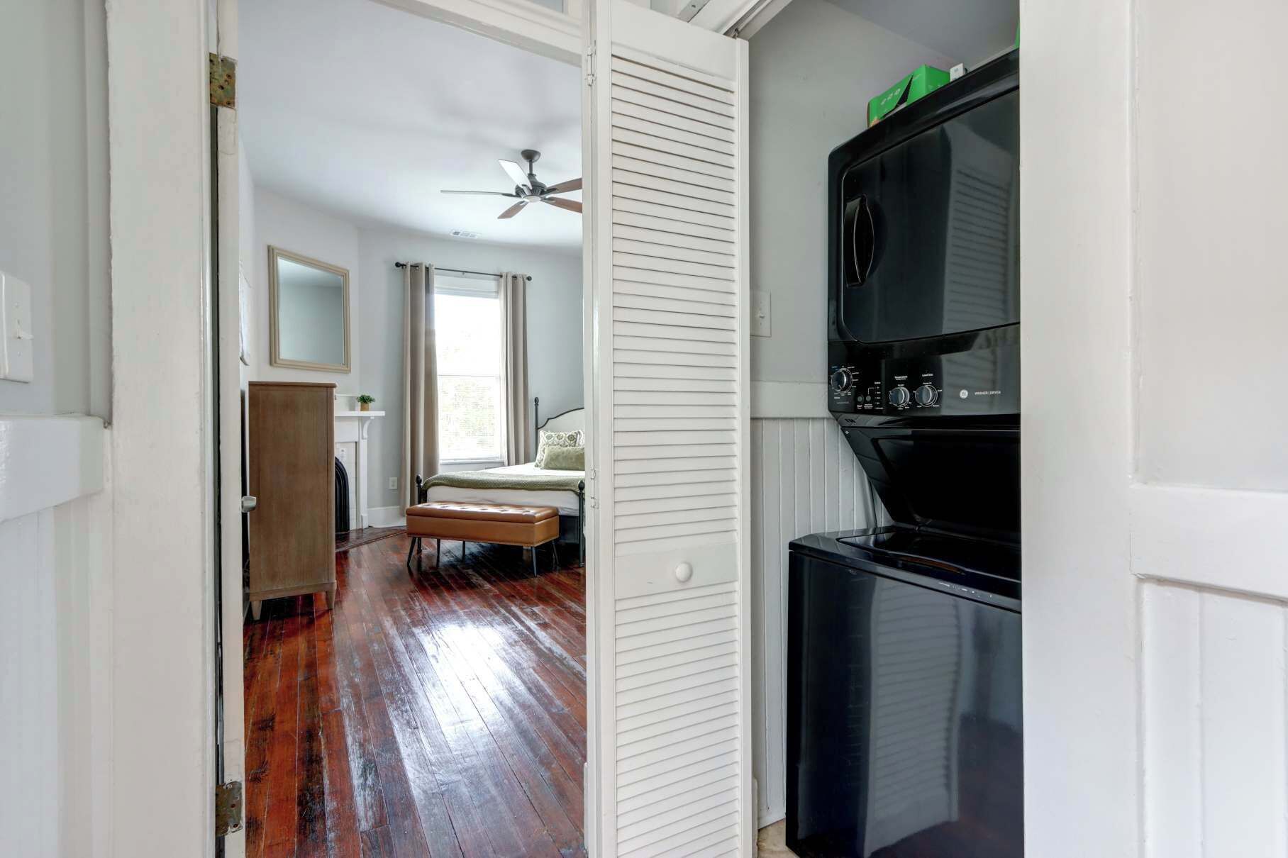 A narrow hallway with a wooden floor leading to a bedroom with a bed and a window. A black refrigerator is visible on the right side.