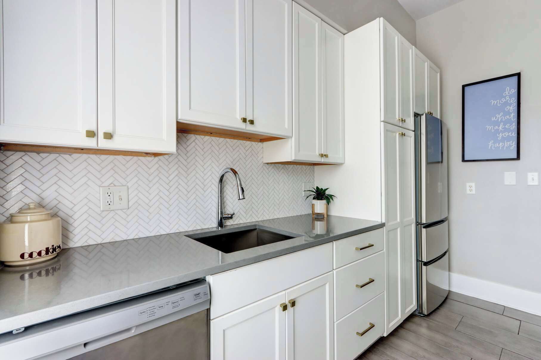 A modern kitchen with white cabinetry, a herringbone tile backsplash, a stainless steel refrigerator, and a dishwasher.