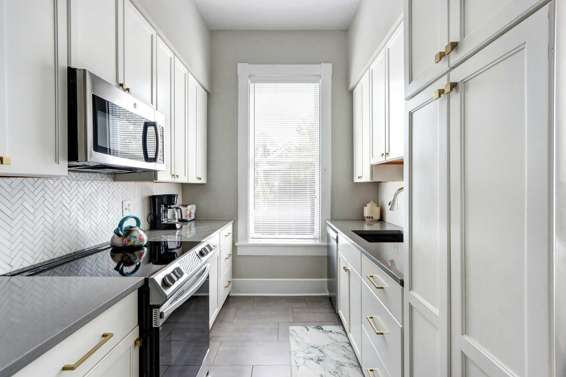 A narrow kitchen with white cabinetry, a microwave, a stove, and a window with blinds.