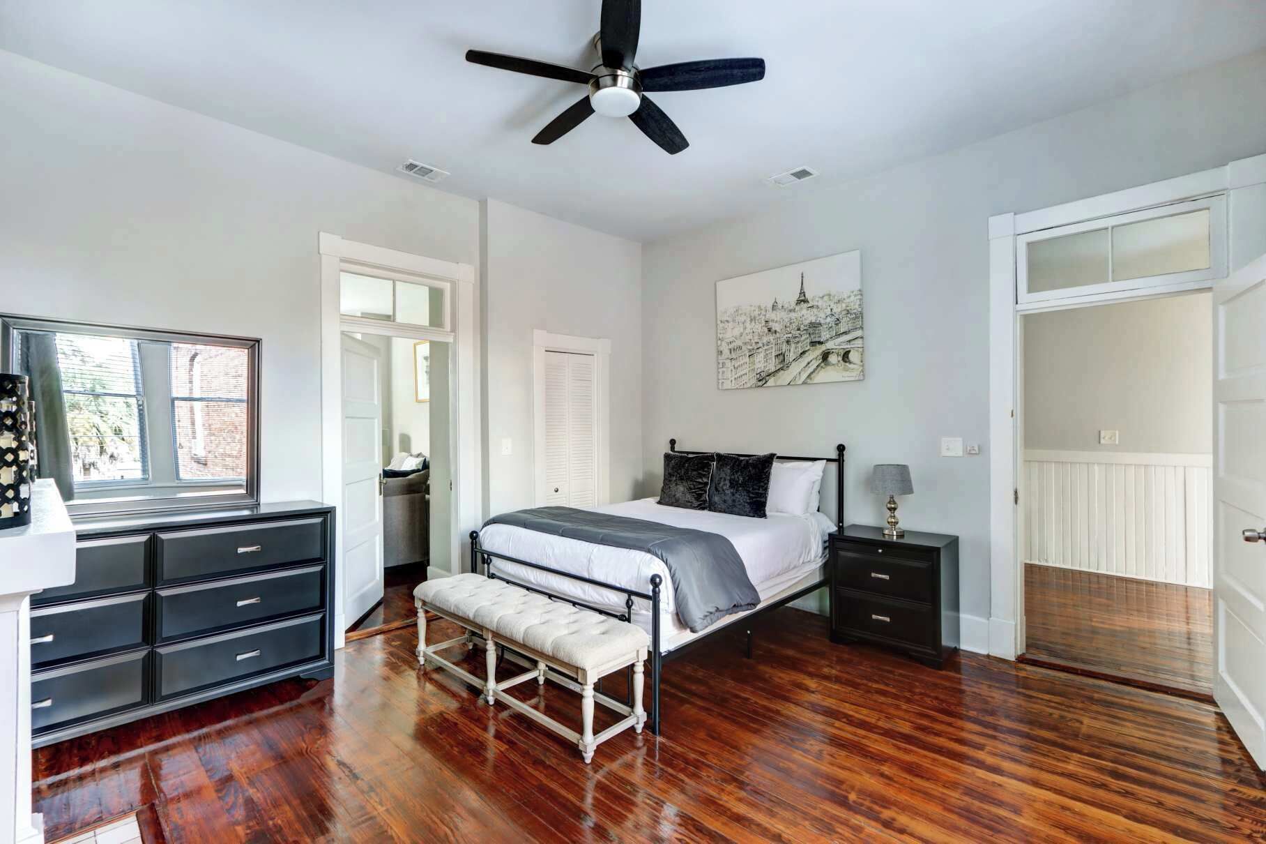 Une chambre à coucher comprenant un lit, une table de chevet, une commode, un banc, un ventilateur de plafond, une fenêtre et une porte.