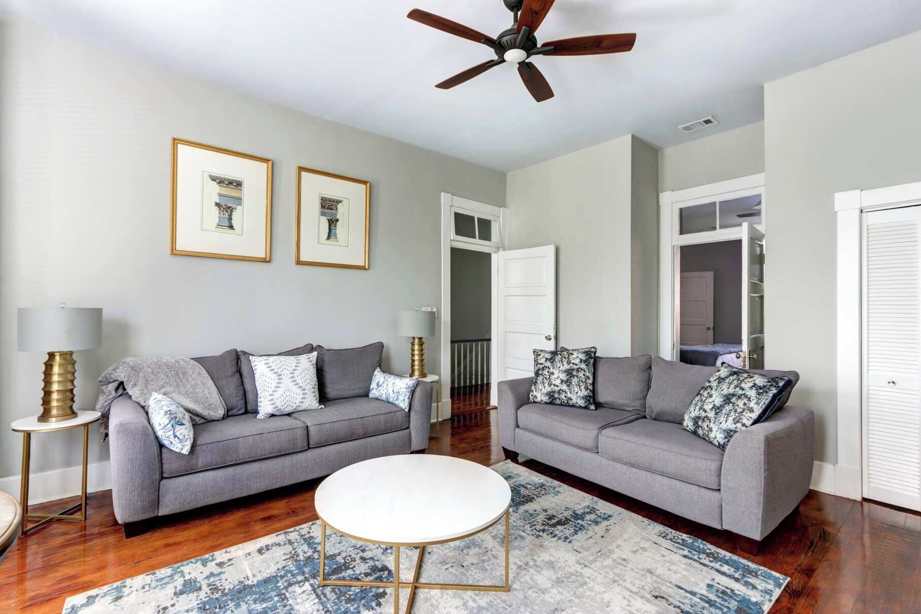 A living room with two grey sofas, a white coffee table, and a patterned rug. There are framed pictures on the wall, a ceiling fan, and a door leading to a closet.
