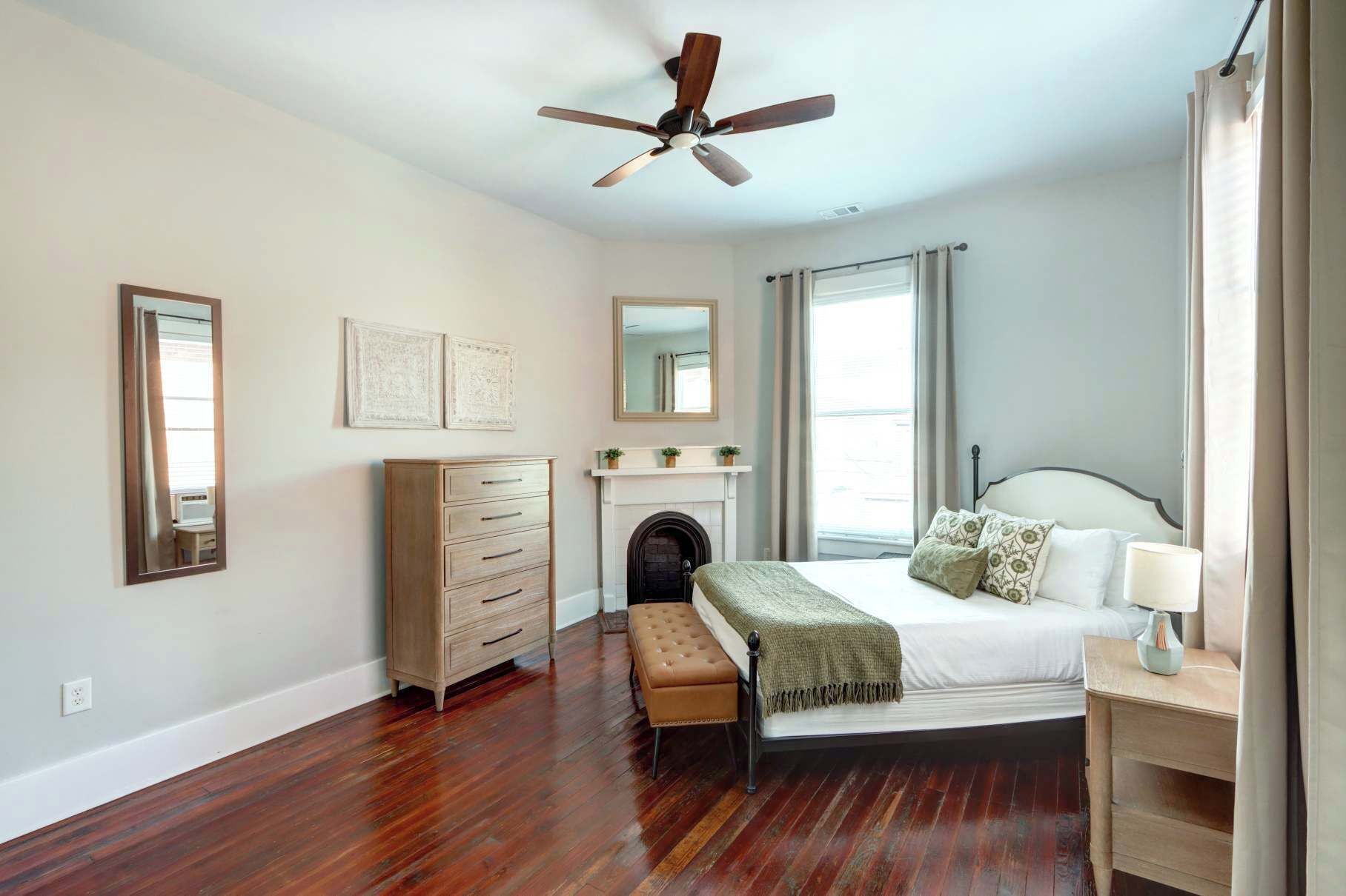 A bedroom with a wooden floor, a bed with a headboard, a nightstand, a dresser, a mirror, a ceiling fan, and a fireplace.