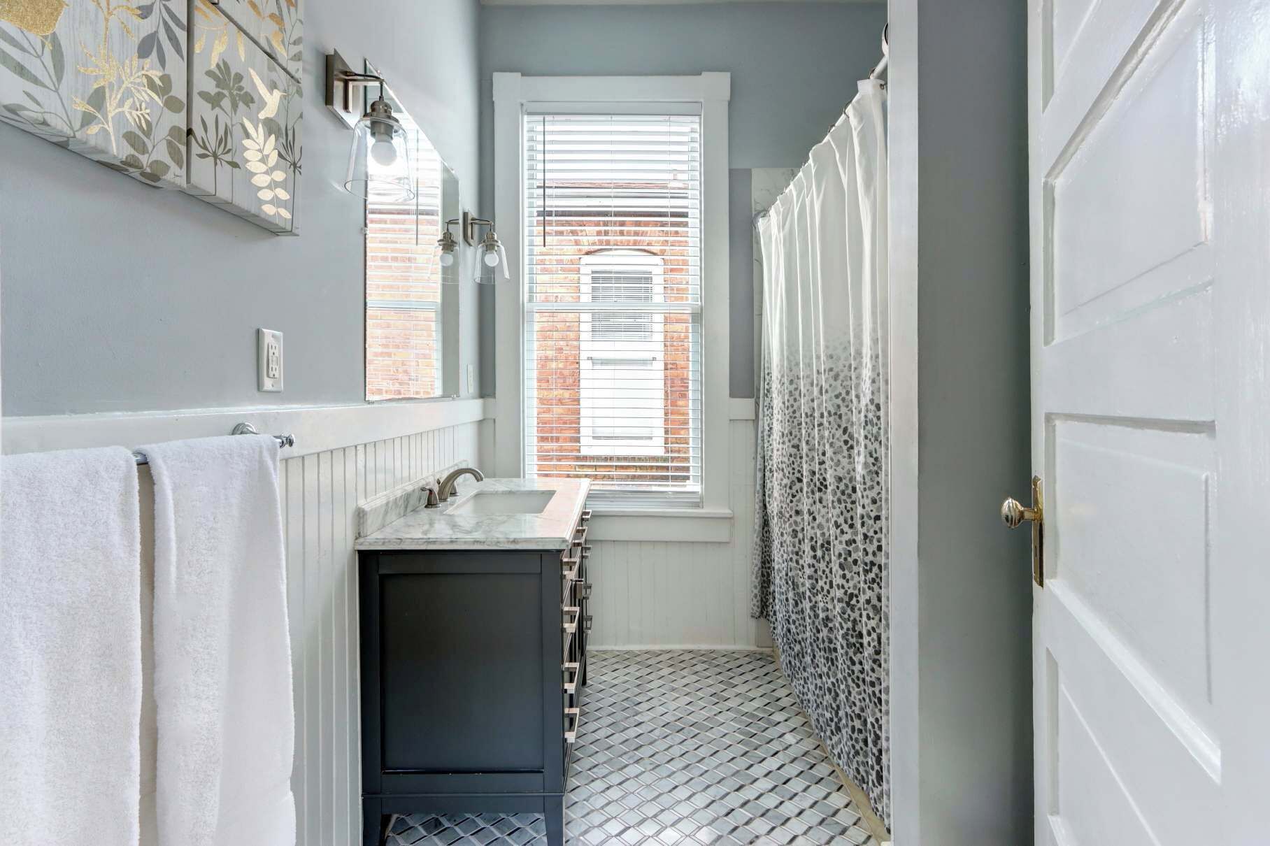 A narrow bathroom with a white sink, a mirror, and a window with blinds.