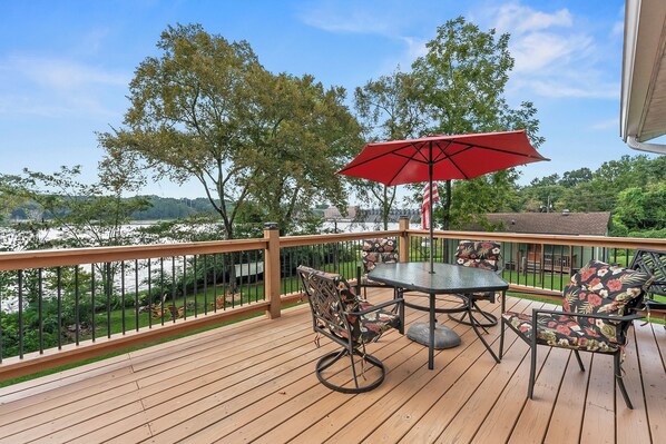 A wooden deck with a table and chairs, a red umbrella, and a view of a body of water and trees.