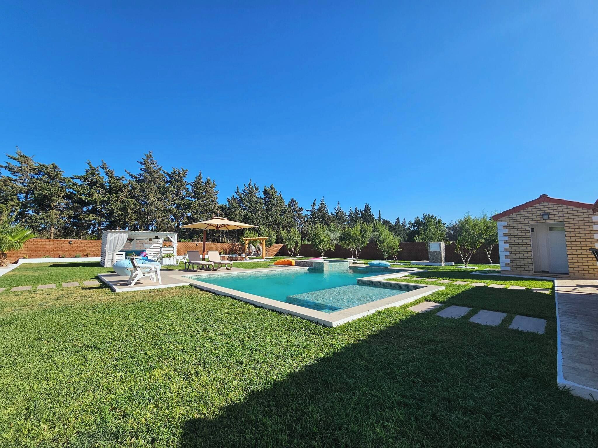 Un jardin avec une piscine, un petit abri de jardin et un espace patio aménagé avec des chaises et des parasols.