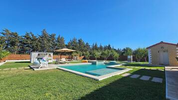 Un jardin avec une piscine, un petit abri de jardin et un espace patio aménagé avec des chaises et des parasols.