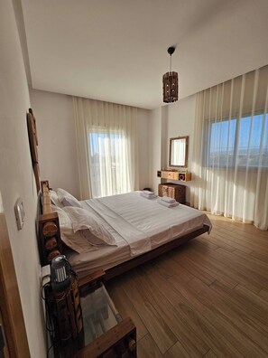 Quarto com cama grande, cabeceira de madeira, mesa de cabeceira com abajur, espelho e janela com cortinas.