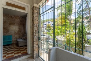 Una habitación con suelo a cuadros, pared de piedra, una verja verde y vistas a una casa y árboles.