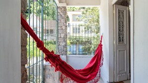 A red hammock hanging between a stone pillar and a metal gate, with a view of a building and greenery in the background. - Casa p 8 Pessoas c Varanda a 400m Museu Imperial (Petrópolis)