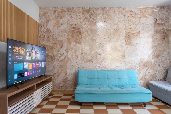 Un salon avec un téléviseur posé sur un meuble en bois, un canapé bleu et un sol à carreaux.