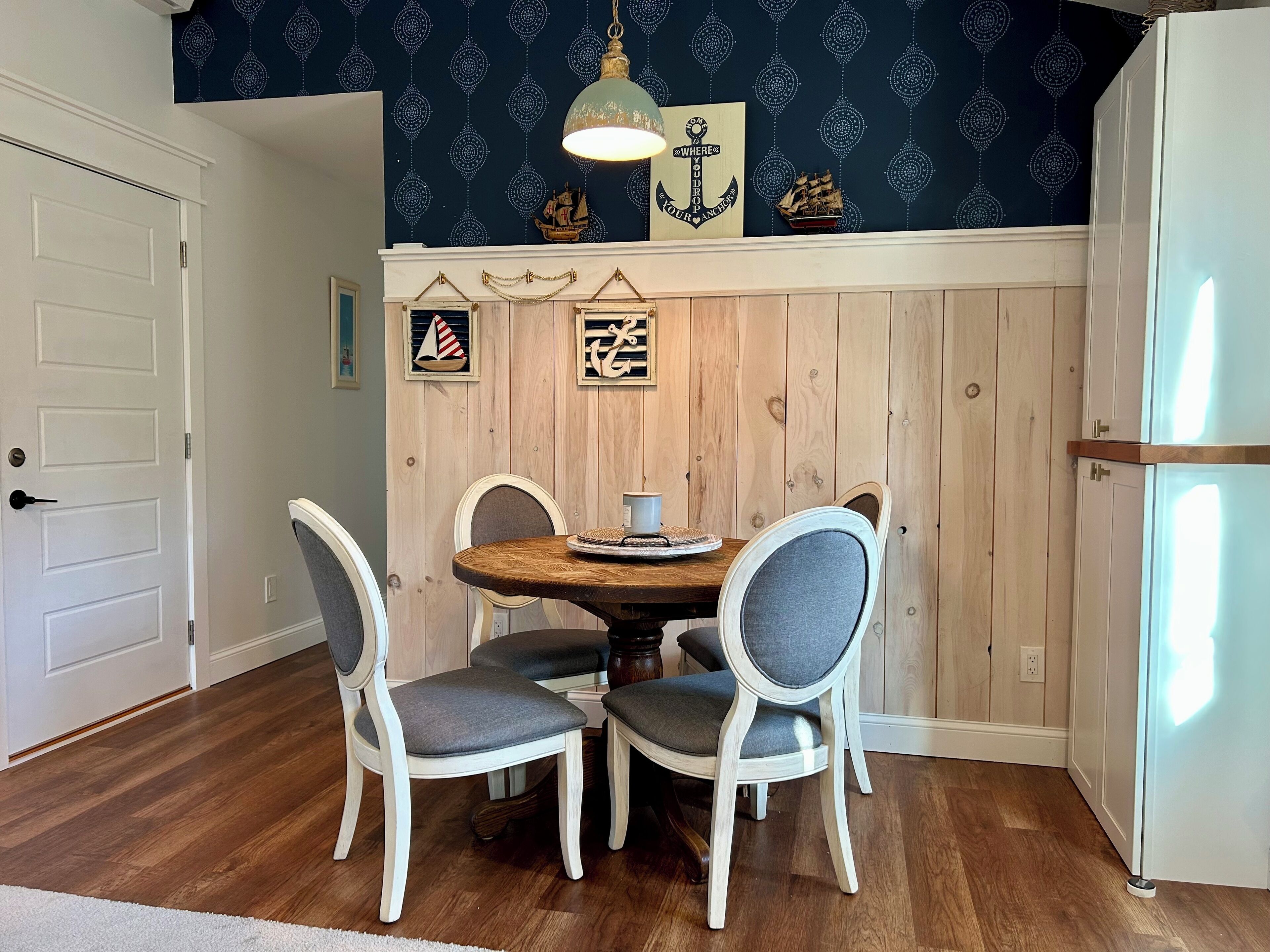 Un coin repas avec une table en bois et quatre chaises, un mur décoratif orné d’œuvres d’art sur le thème de la marine, et un luminaire suspendu.