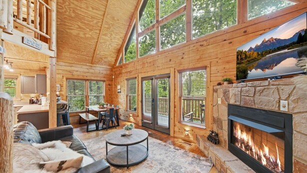 A log cabin interior with a fireplace, large windows, a dining area, and a cozy seating arrangement. - Little Woods Cabin | Family-Friendly w/ Games! (Sevierville)