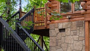 A wooden cabin with a stone wall, a staircase, and a sign indicating Staycation services. - Little Woods Cabin | Family-Friendly w/ Games! (Sevierville)
