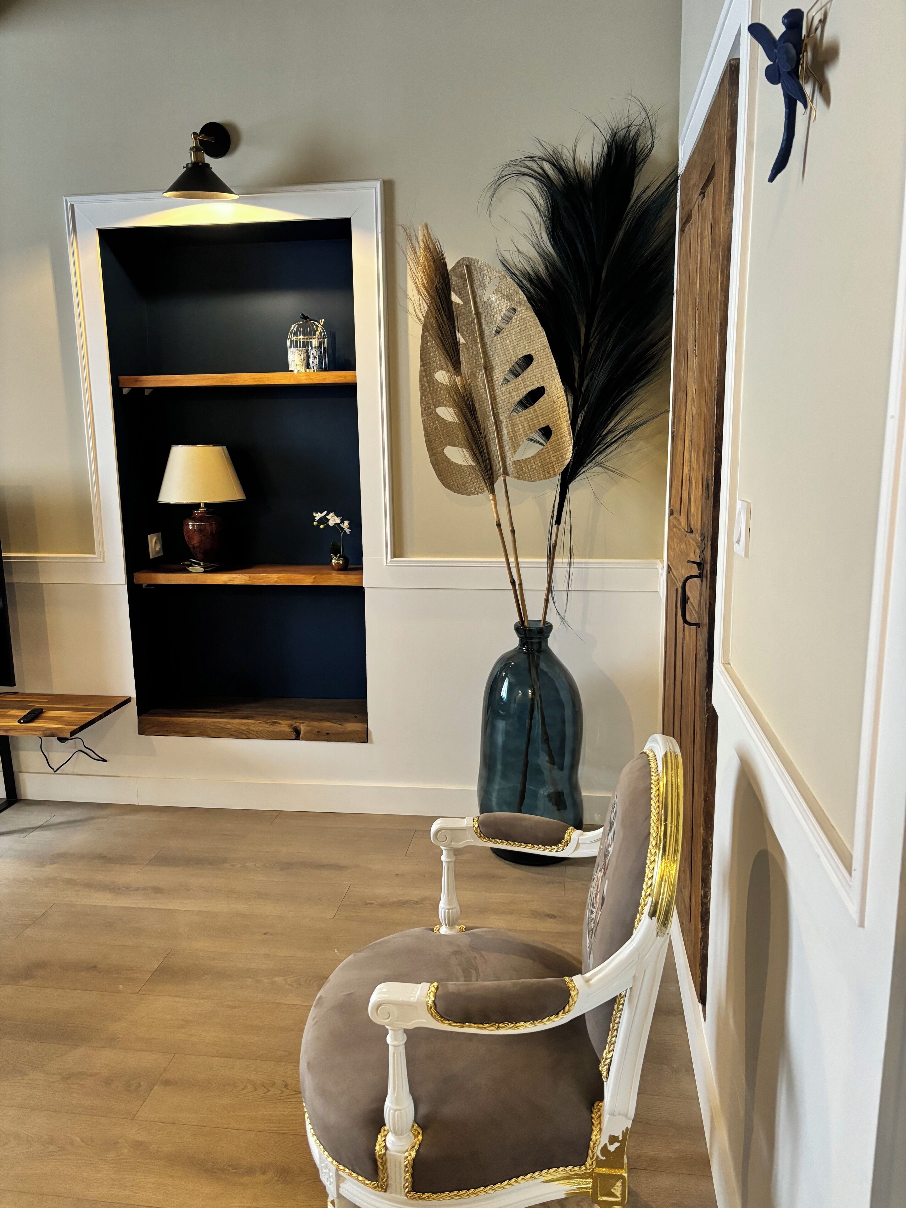 A room with a wooden floor, a white chair with gold trim, a dark blue bookshelf, a vase with decorative leaves, and a wall-mounted fan.