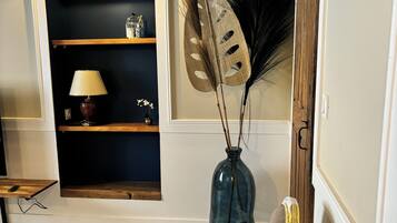 A room with a wooden floor, a white chair with gold trim, a dark blue bookshelf, a vase with decorative leaves, and a wall-mounted fan.
