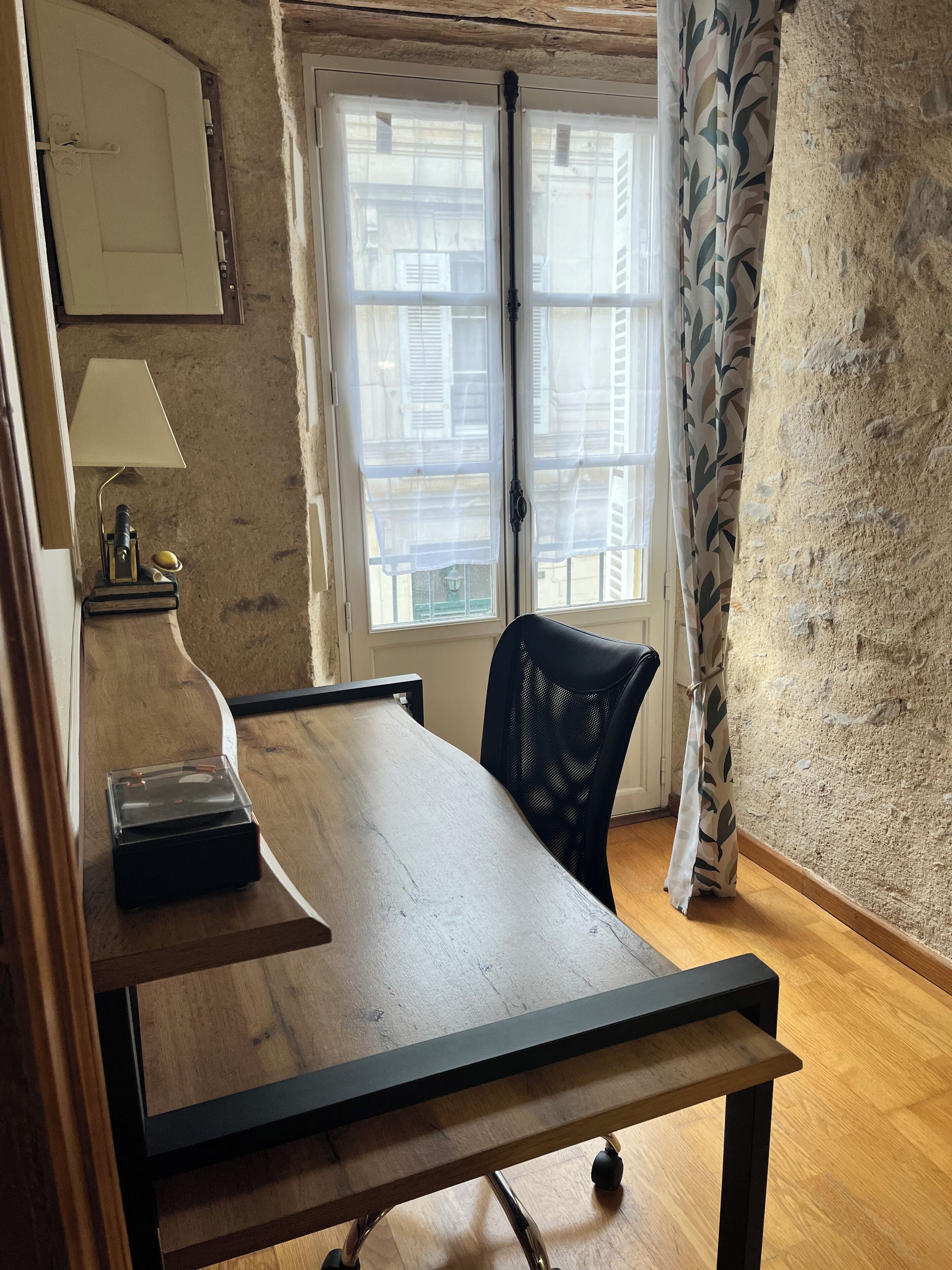 Uma mesa de madeira com uma luminária, uma cadeira e uma janela com cortinas leves.