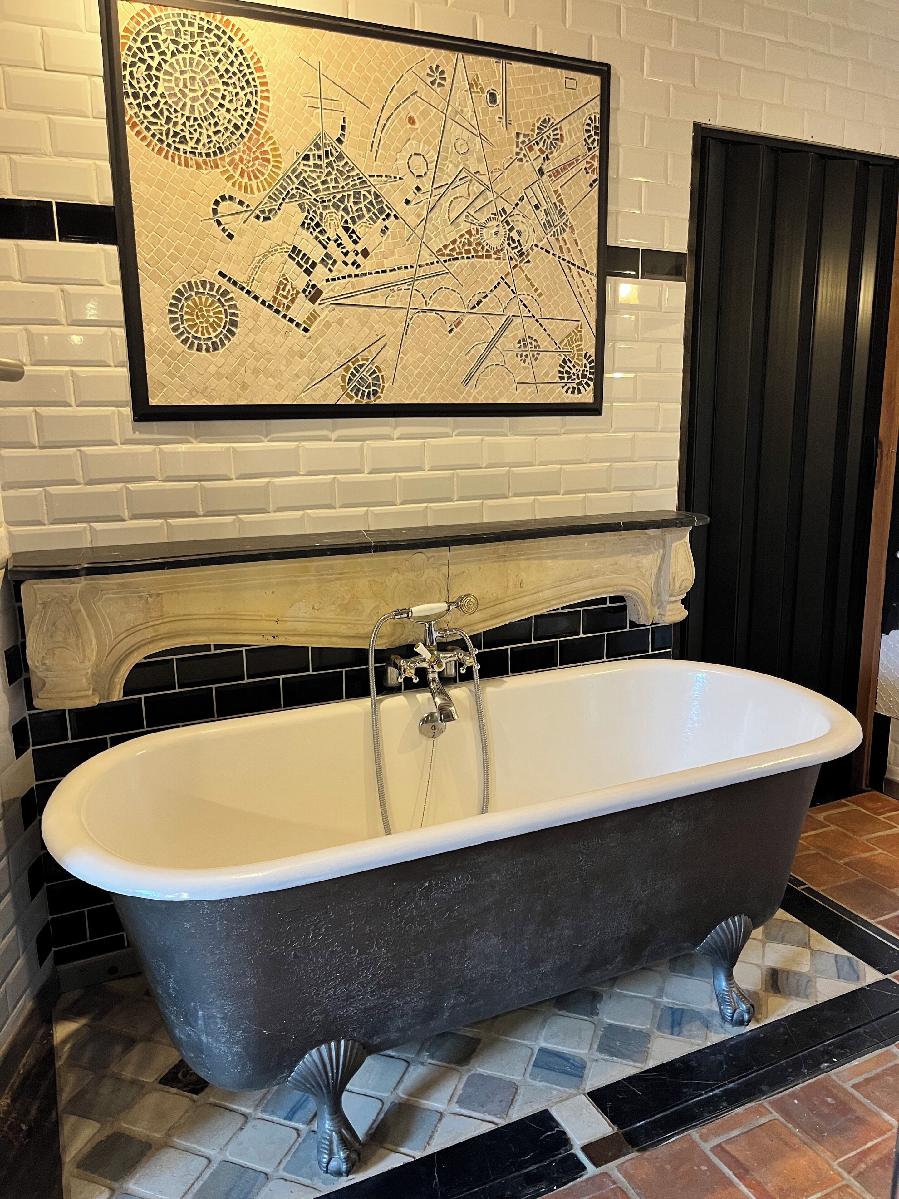 Une salle de bain avec une baignoire sur pieds, des carreaux de mosaïque noirs et blancs, et une œuvre d’art abstraite encadrée au-dessus de la baignoire.
