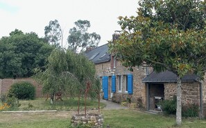 Een stenen huis met blauwe luiken, een tuin met een boom en een stenen muur op de achtergrond.
