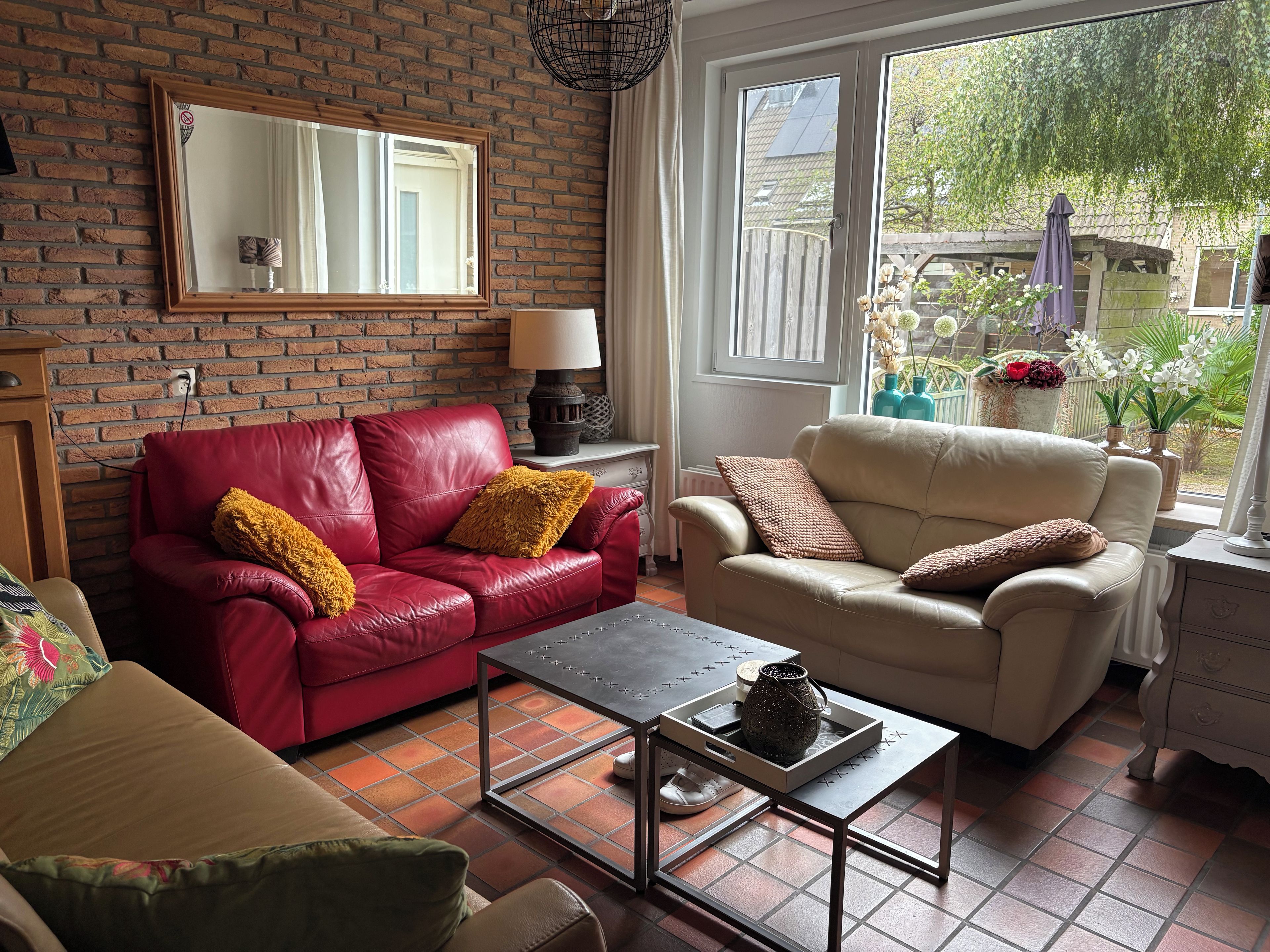 Sala de estar com sofá de couro vermelho, sofá bege e mesa de centro. Há uma parede de tijolos e uma janela com vista para o exterior.